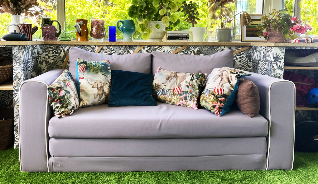 plantes vertes tablettes en pierre naturelle et brute revêtement de sol gazon coussins colorés murs bleus bibelots vases objets décoratifs livres large ouverture sur le jardin vitrages sur les trois côtés de la véranda pièce de transition entrée salle de jeux vestiaire